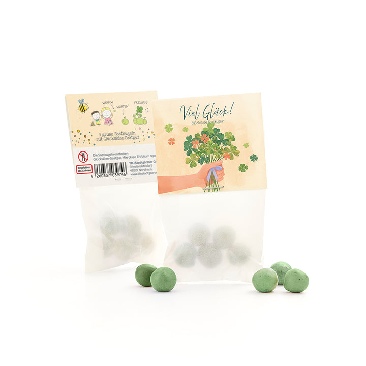 Bolsa con bolas de semillas verdes y semillas de trébol de la suerte, junto a varias bolas verdes sueltas, en el embalaje un motivo de felicitación y una ilustración de trébol.