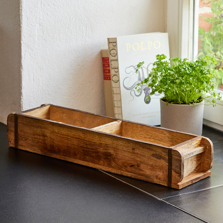 Caja de madera con tres compartimentos sobre la encimera de la cocina, junto a ella un libro de cocina y un bote con hierbas frescas.