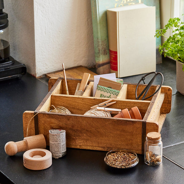 Caja de madera con herramientas de jardinería, incluidos paquetes de semillas, pala, tijeras, pequeñas macetas de barro, cordel y frasco de hierbas, sobre una encimera de cocina.
