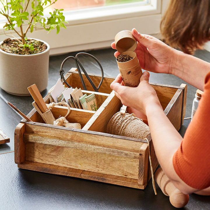Persona llenando una maceta biodegradable con tierra sobre una caja de madera con accesorios de jardinería.