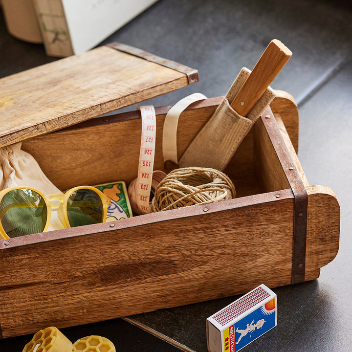 Caja de madera que contiene varios artículos, incluidas gafas de sol amarillas, una cinta métrica, un carrete de cuerda, una caja de cerillas y una herramienta con mango de madera.