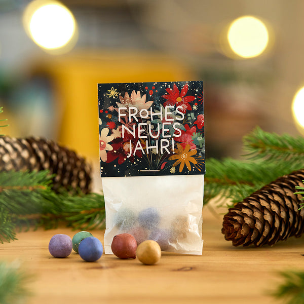 Una bolsa de adornos de colores y una tarjeta que dice "¡Feliz Año Nuevo!" descansan sobre una mesa de madera, rodeadas de ramas de abeto y piñas.