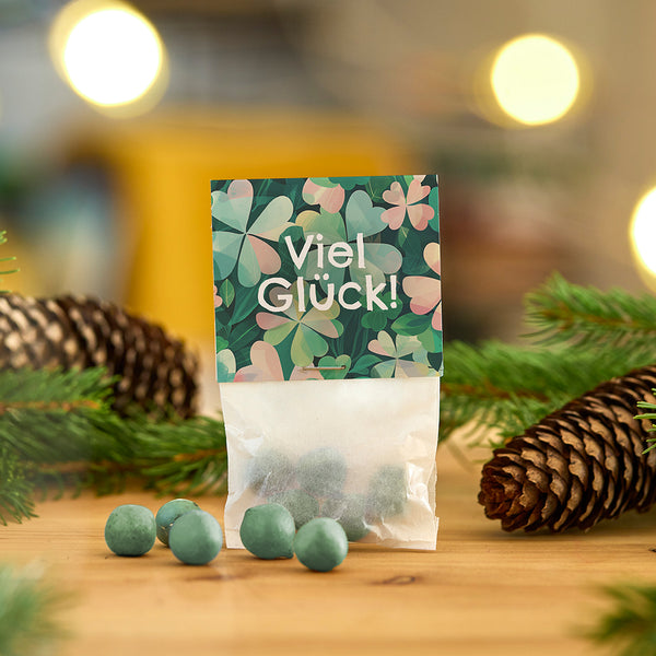 Bolsas con bolas verdes y la inscripción "¡Buena suerte!" delante de adornos navideños con ramas de abeto y piñas.