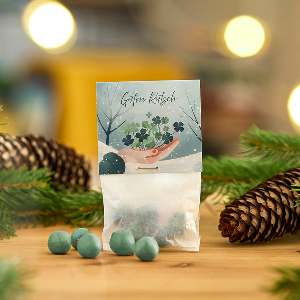 Bolsa con bolas verdes y tarjeta con la inscripción “Feliz Año Nuevo”, rodeada de piñas y ramas.