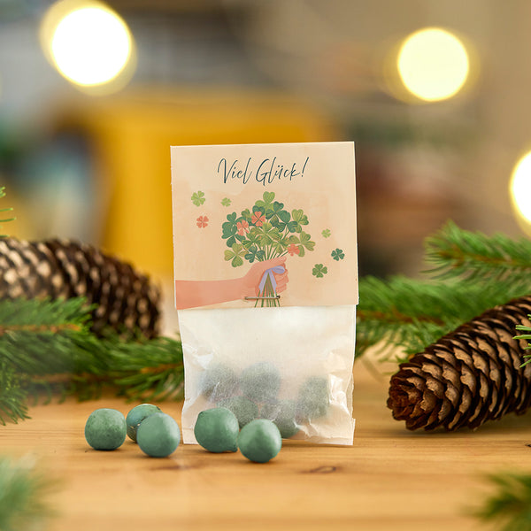 Bolsa de bolas de Navidad de color verde, decorada con una tarjeta que dice "¡Buena suerte!" y un ramo de tréboles rodeado de ramas de abeto y piñas.