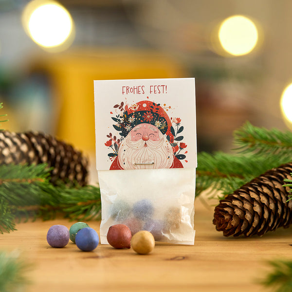 Bolsa con bolas de colores, tarjeta navideña con motivo de Papá Noel y la inscripción “¡Feliz Navidad!”, piñas y ramas de fondo.