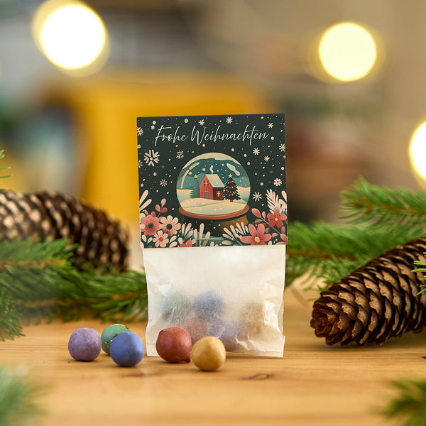 Bolsa con bolas de colores y una tarjeta navideña con la inscripción "Feliz Navidad" delante de adornos navideños con piñas y ramas.
