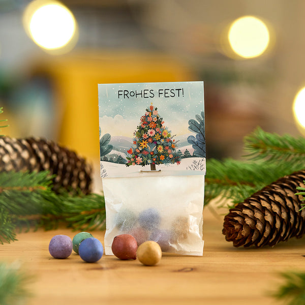 Bolsa de adornos de colores delante de una tarjeta con un árbol de Navidad y las palabras "Feliz Navidad", rodeado de ramas de abeto y una piña.