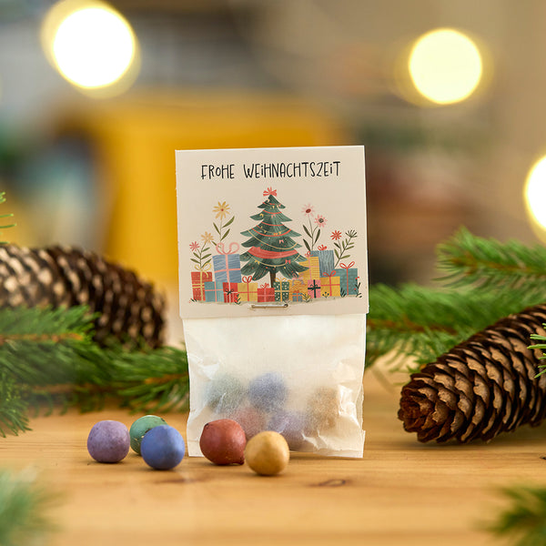 Una pequeña bolsa de adornos de colores, decorada con una tarjeta navideña que dice "Feliz Navidad", rodeada de ramas de abeto y una piña.