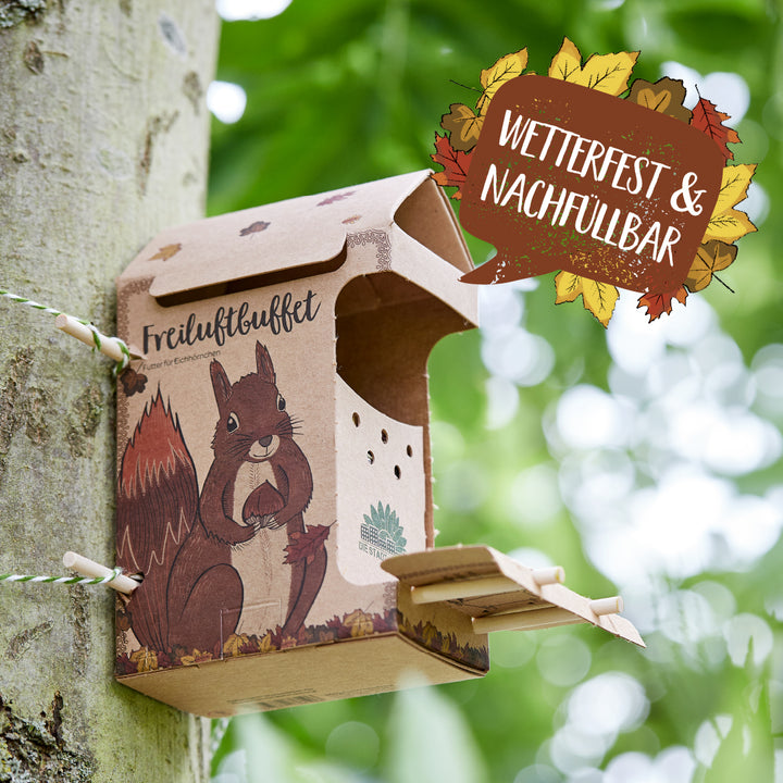 Comedero de cartón con motivo de ardilla fijado a un árbol, con la inscripción "Buffet al aire libre" y un cartel que dice "Resistente a la intemperie y rellenable".