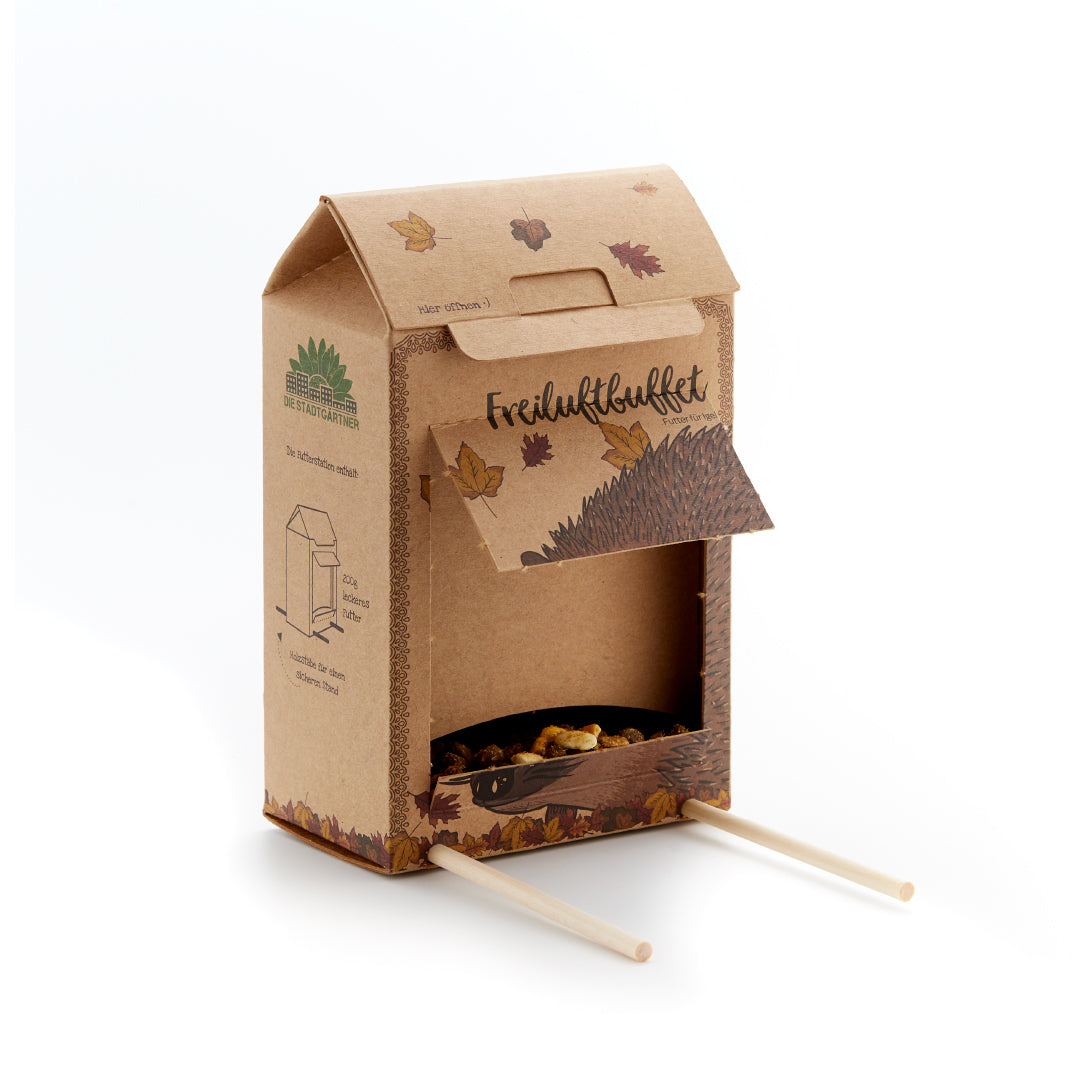 Caja de cartón con solapa abierta, hojas de otoño, ilustración de erizo y dos palos de madera, sirve como comedero para erizos.