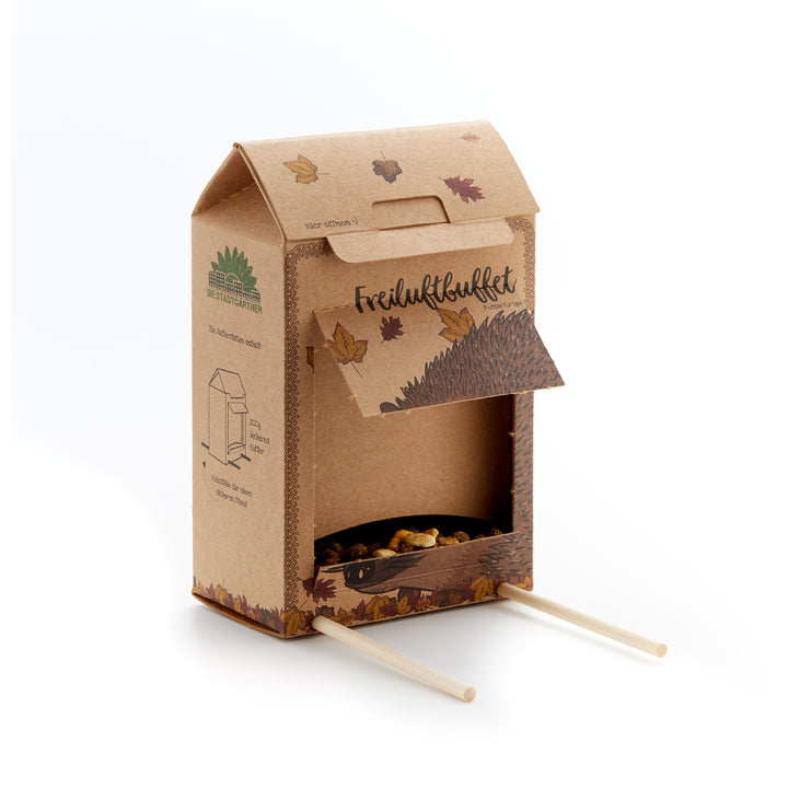 Caja de cartón con solapa abierta, hojas de otoño, ilustración de erizo y dos palos de madera, sirve como comedero para erizos.