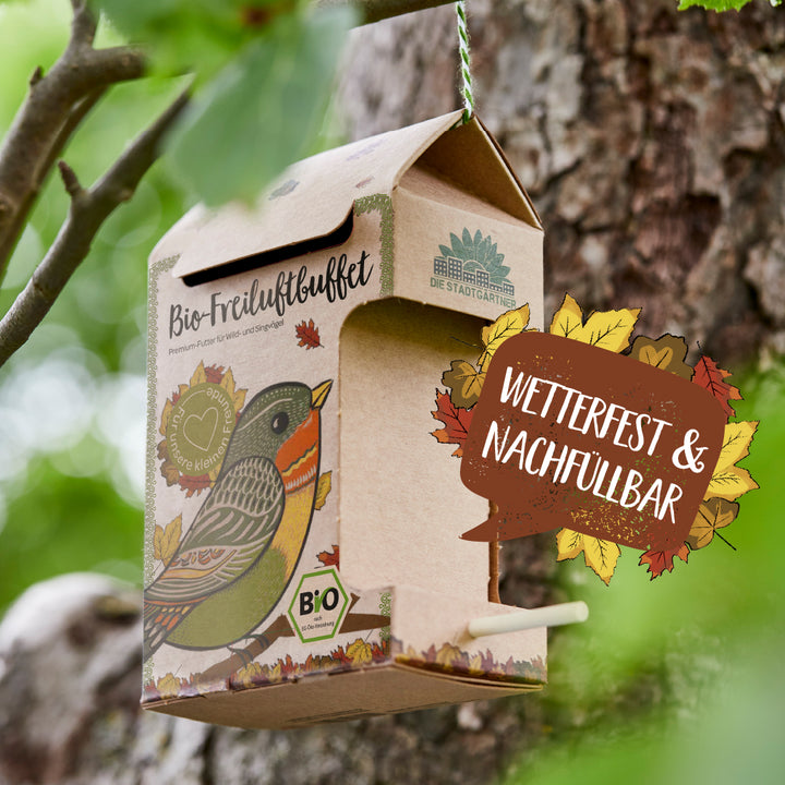 Comedero para pájaros de cartón con ilustración de pájaro y la inscripción “Buffet orgánico al aire libre” colgado en un árbol, junto a él el texto “Resistente a la intemperie y rellenable”.