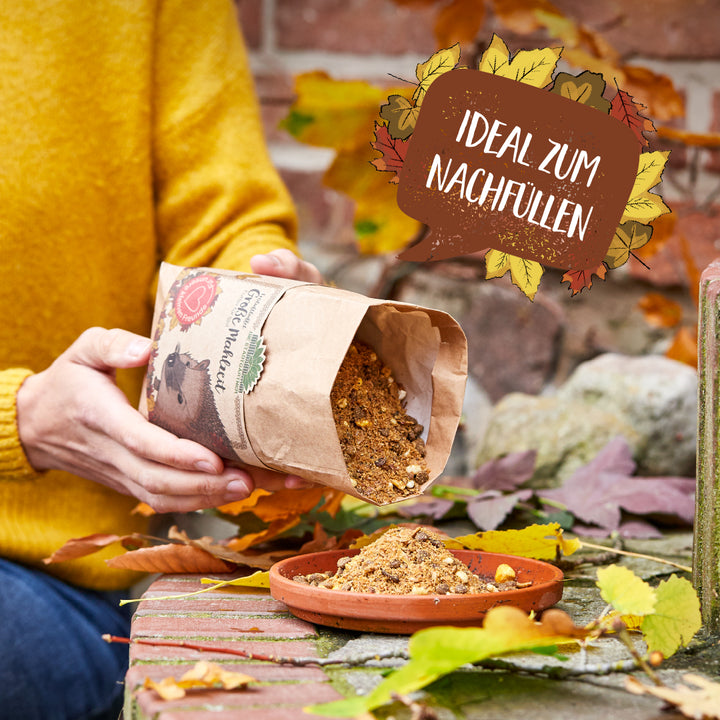 Una persona con un suéter amarillo vierte comida para erizos desde una bolsa de papel a un recipiente poco profundo rodeado de hojas de otoño; al lado hay un cartel que dice "Ideal para rellenar".