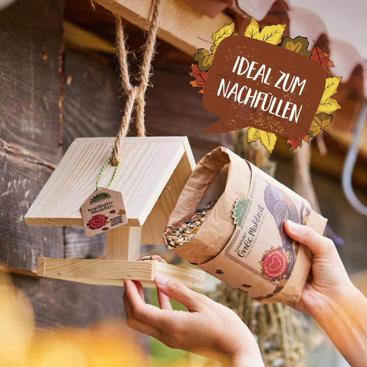 Una persona llena una pajarera de madera con alpiste de una bolsa de papel; en la imagen se lee "Ideal para rellenar".