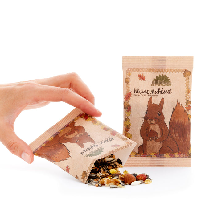Una mano sostiene una bolsa de papel con comida para ardillas, de la que caen semillas y nueces sobre una superficie blanca, junto a otra bolsa con una ilustración de una ardilla.