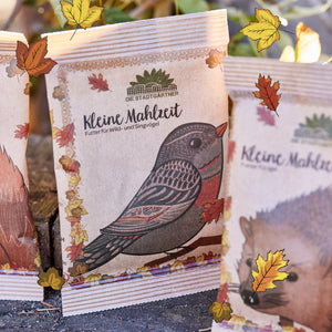 Bolsa de papel con estampado de pájaros y la inscripción "Small Meal - Food for Wild and Songbirds" de Die Stadtgärtner, rodeada de hojas otoñales.