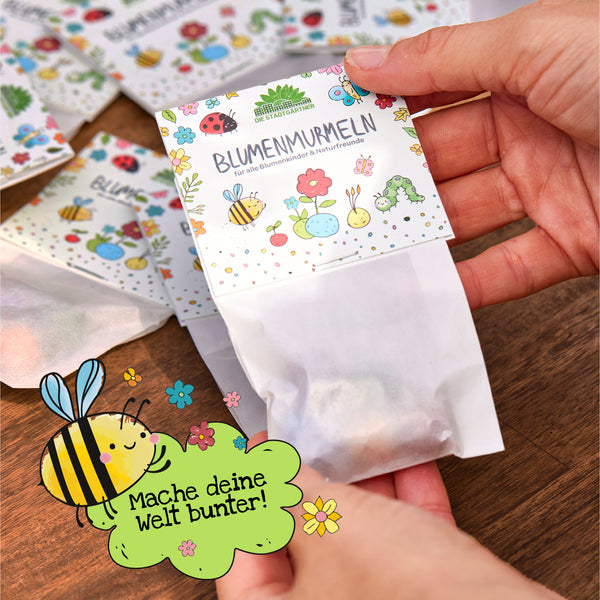 Una mano sostiene un paquete de semillas de colores con la etiqueta "Canicas de flores" sobre una mesa de madera; en primer plano hay un dibujo de una abeja con el texto "¡Haz que tu mundo sea más colorido!"
