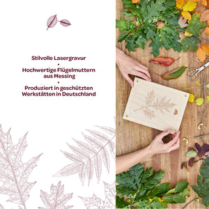 Dos manos sostienen una placa de madera con un delicado grabado láser de una hoja, rodeada de varias hojas de otoño coloridas y materiales de artesanía; junto a ella hay un texto con información sobre el grabado láser, tuercas de mariposa de latón y producción en Alemania.