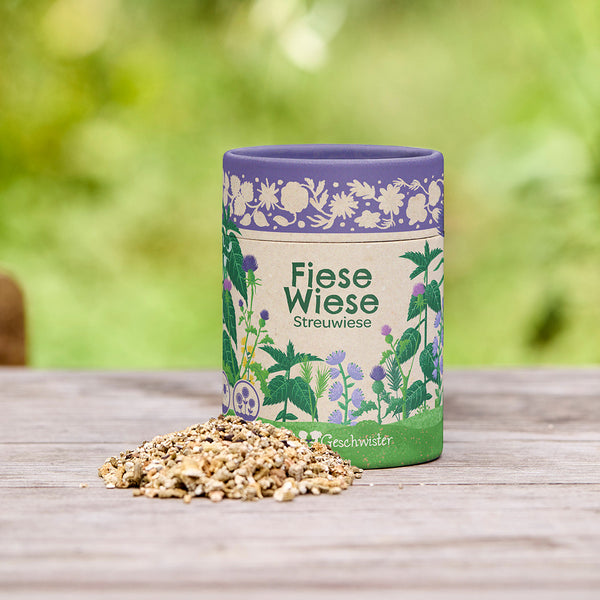 Lata etiquetada como "Fiese Wiese Streuwiese" (Prado disperso de Fiese Wiese) con una mezcla de semillas en una mesa de madera al aire libre.