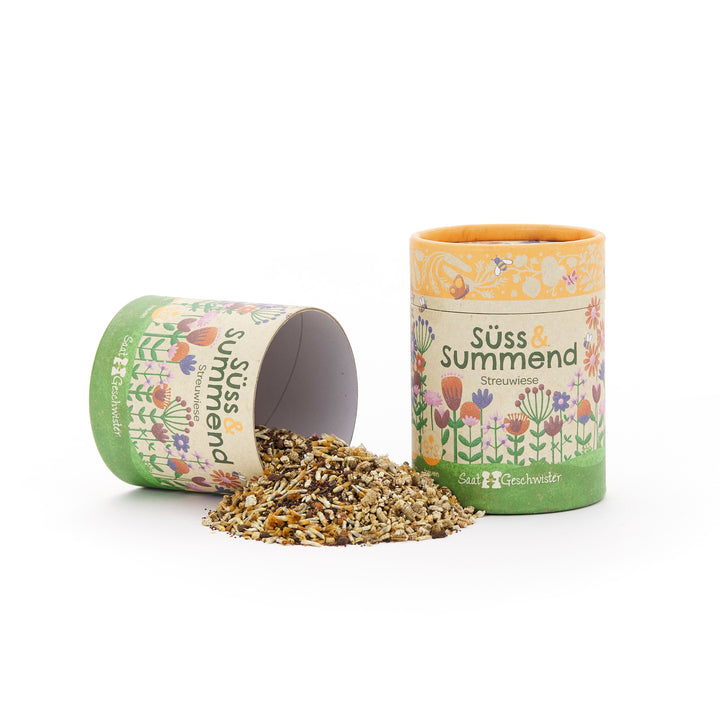 Dos cajas de cartón de colores con motivos florales, una vertical y otra acostada, con semillas de flores cayendo de ellas, etiquetadas como “Sweet & Buzzing Meadow”.