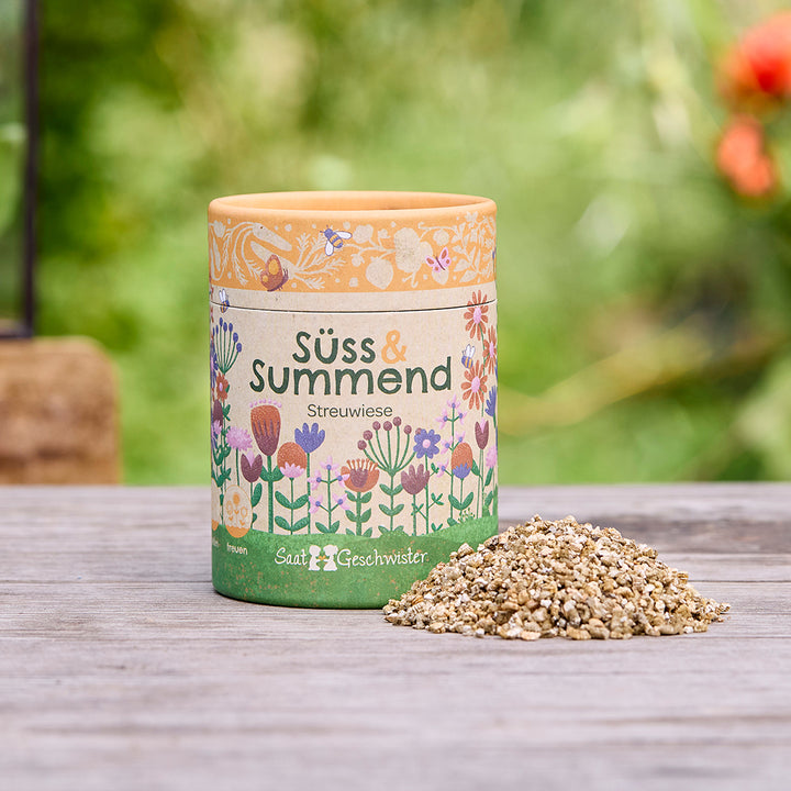 Lata redonda con la inscripción “Sweet & Buzzing Meadow” y un motivo floral, junto a ella un pequeño montón de semillas sobre una mesa de madera, fondo verde difuminado.