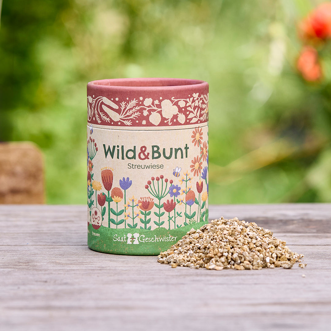 Una lata con la inscripción "Wild & Bunt Streuwiese" y motivos florales ilustrativos, junto a un pequeño montón de semillas, sobre una mesa de madera al aire libre.