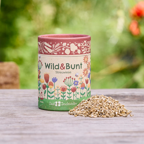 Una lata con la inscripción "Wild & Bunt Streuwiese" y motivos florales ilustrativos, junto a un pequeño montón de semillas, sobre una mesa de madera al aire libre.