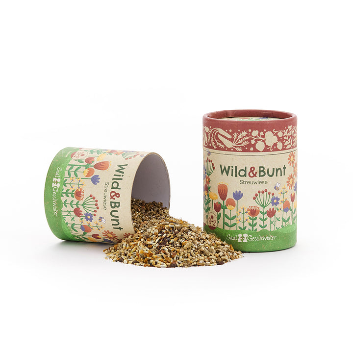 Dos latas de colores con la inscripción "Wild & Colorful Streuwiese" (Esparcimiento salvaje y colorido en la pradera), una de ellas abierta, de la que caen semillas sobre la mesa.