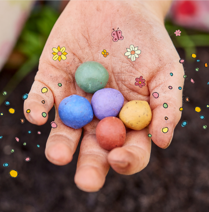 Una mano sostiene cinco bombas de semillas de colores, rodeadas de flores dibujadas, puntos y una mariposa.