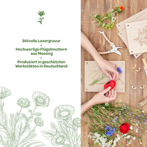 Dos manos colocan flores de colores sobre un marco de madera; junto a él, sobre una mesa de madera, se encuentran herramientas, tuercas de mariposa de latón y placas de madera grabadas. A la izquierda, texto con información sobre el elegante grabado láser y su producción en Alemania.