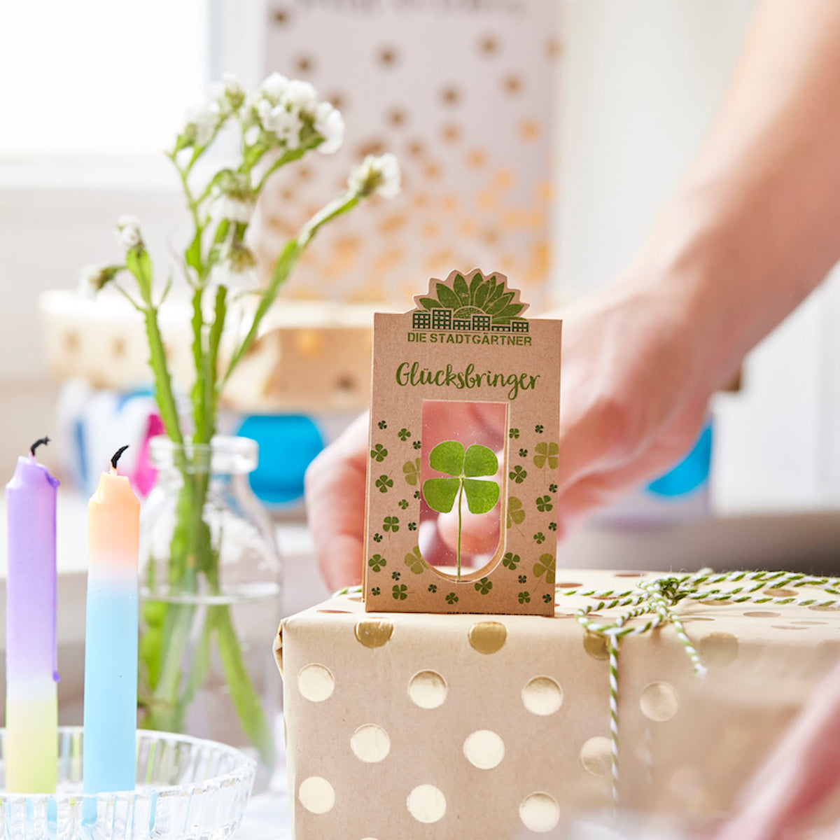 Una mano sostiene un regalo envuelto con un trébol de cuatro hojas y las palabras "Lucky Charm" encima, con flores y velas de colores visibles en el fondo.