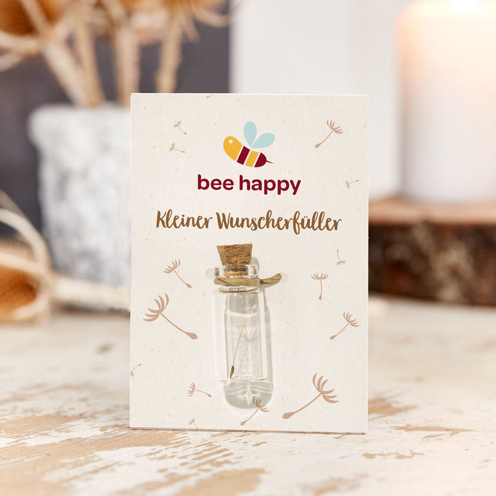 Pequeña botella de cristal con corcho sobre una tarjeta con la inscripción “abeja feliz, pequeña cumplidora de deseos”, decorada con motivos de diente de león.