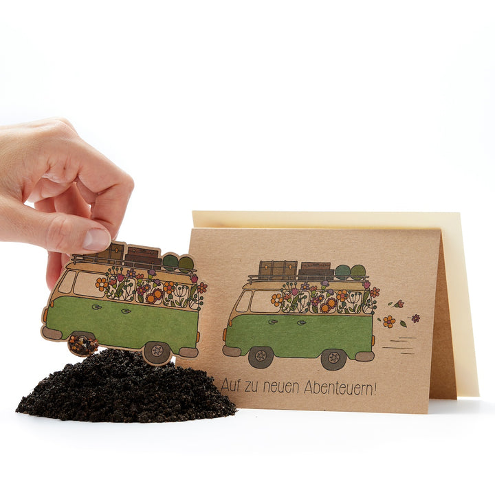 Hand pega en la tierra una tarjeta verde con forma de furgoneta y un motivo de flores, y junto a ella una tarjeta de felicitación con el texto “¡Hacia nuevas aventuras!”.