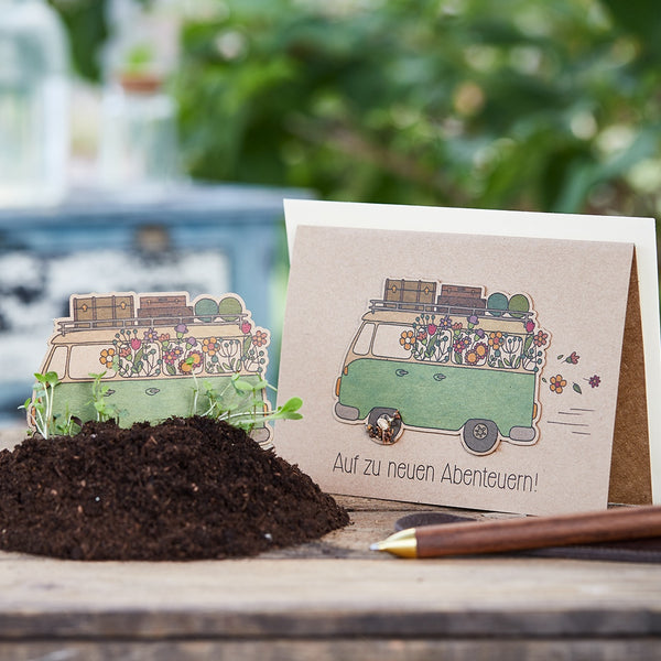 Tarjeta de felicitación con un autobús verde lleno de flores y equipaje, al lado tierra con pequeñas plantas, texto en la tarjeta: "¡Hacia nuevas aventuras!"