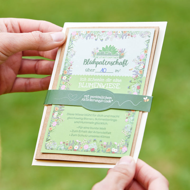 Persona sosteniendo una tarjeta con la inscripción "Blühpateenschaft" y una descripción de un regalo en forma de un prado de flores.