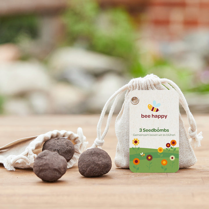 Bombas de semillas junto a una bolsa etiquetada "Bee Happy 3 Seed Bombs" sobre una mesa de madera con un fondo verde borroso.