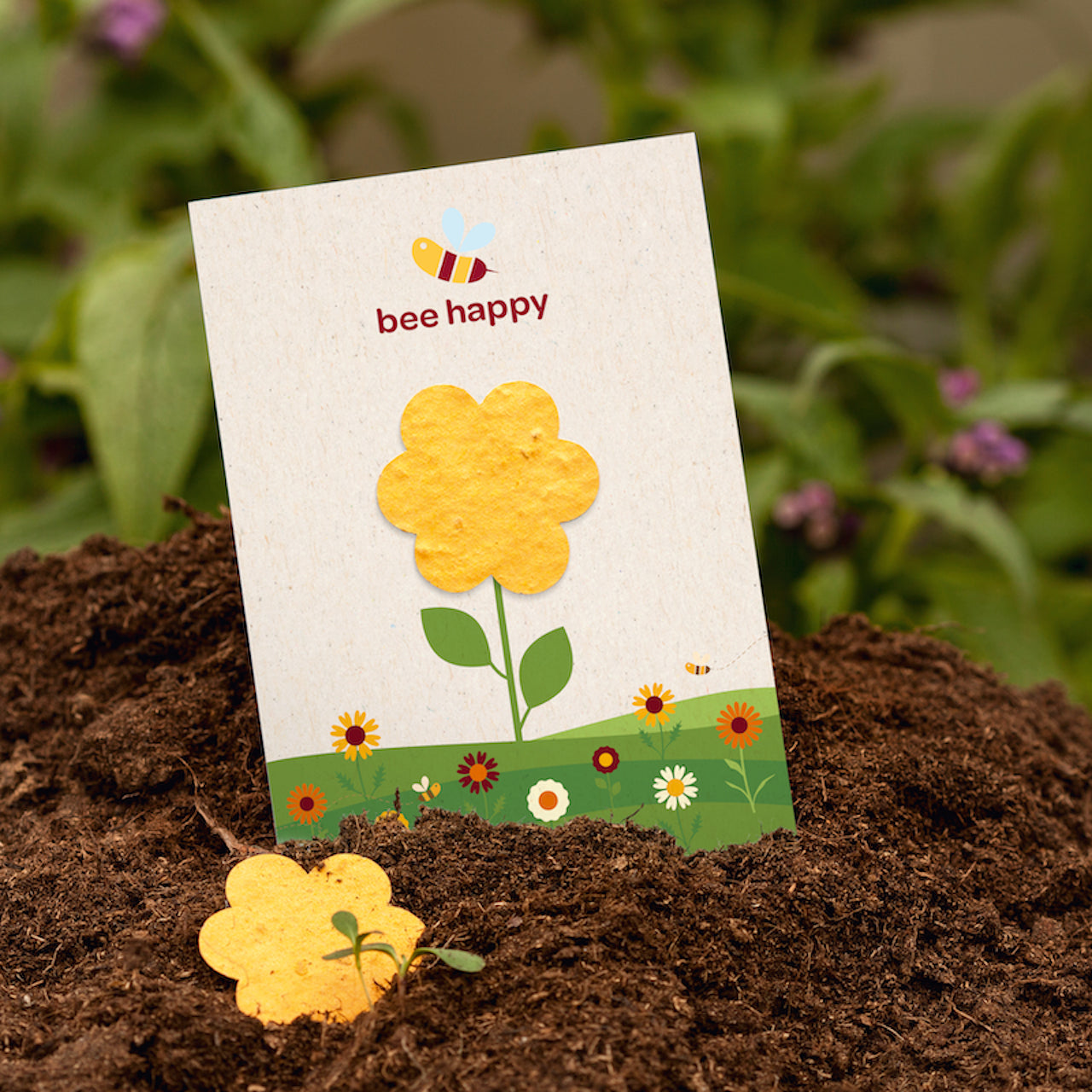 Una tarjeta con una flor amarilla y la inscripción "abeja feliz" está pegada en la tierra; en primer plano hay una flor de papel amarilla; en el fondo hay plantas verdes.