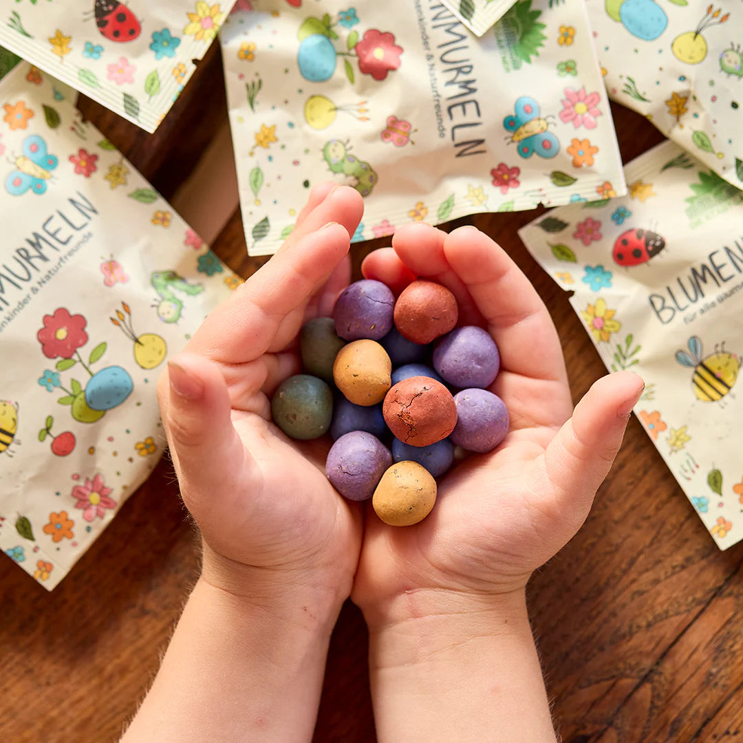 Las manos de los niños sostienen bombas de semillas de colores, mientras que en el fondo se encuentran envases impresos de colores con motivos de flores e insectos.