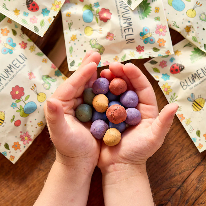 Las manos de los niños sostienen bombas de semillas de colores, mientras que en el fondo se encuentran envases impresos de colores con motivos de flores e insectos.