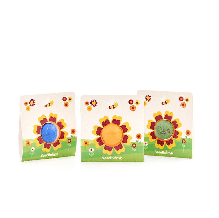 Tres bombas de semillas de colores en un embalaje con motivos de girasoles y abejas sobre fondo blanco.