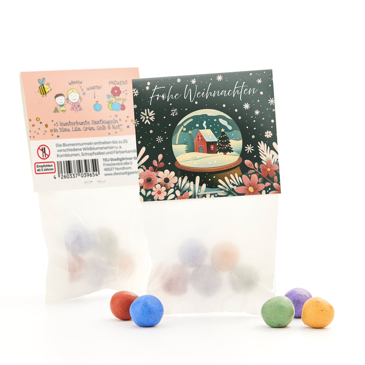 Dos pequeñas bolsas de bolas de semillas de colores, una con una tarjeta de felicitación navideña con un motivo de bola de nieve y la inscripción "Feliz Navidad", al lado bolas de colores sobre un fondo blanco.
