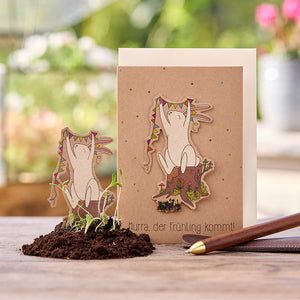 Tarjeta de felicitación con ilustración de un alegre conejito sosteniendo un banderín en el tocón de un árbol; texto: "¡Hurra, se acerca la primavera!"; pequeñas plantas en el suelo en primer plano.