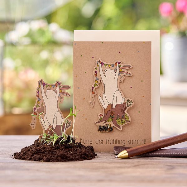 Tarjeta de felicitación con ilustración de un alegre conejito sosteniendo un banderín en el tocón de un árbol; texto: "¡Hurra, se acerca la primavera!"; pequeñas plantas en el suelo en primer plano.