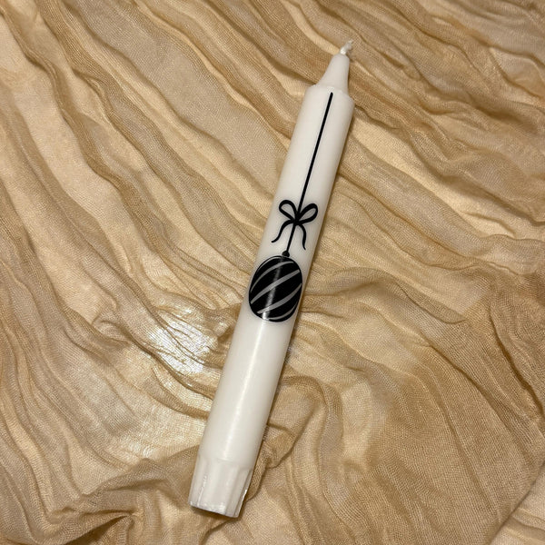 Una vela blanca con un adorno navideño negro como diseño reposa sobre un borde de tela dorada.