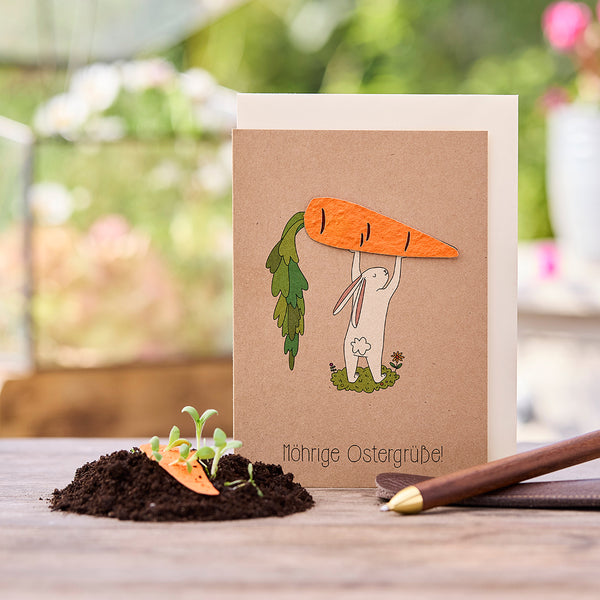 Tarjeta de Pascua con un conejito dibujado sosteniendo una zanahoria grande, al lado tierra con pequeñas plantas y una decoración de zanahoria, texto "¡Saludos de Pascua de zanahoria!".
