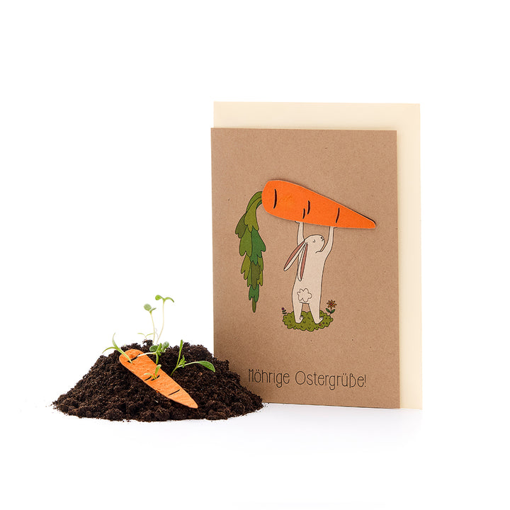 Una tarjeta de Pascua con el dibujo de un conejito sosteniendo una zanahoria grande sobre su cabeza, junto a un pequeño montículo de tierra con una zanahoria de papel y plantas que brotan. El texto dice: "¡Saludos de Pascua con zanahorias!"