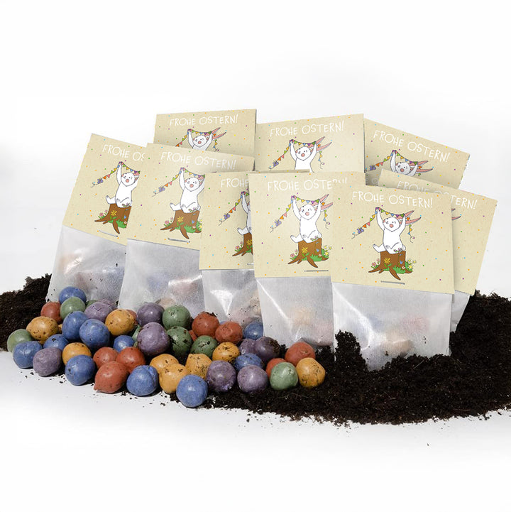 En el suelo se encuentran bombas de semillas de colores y detrás de ellas varias bolsitas con la inscripción “¡Felices Pascuas!” y un motivo de conejito.