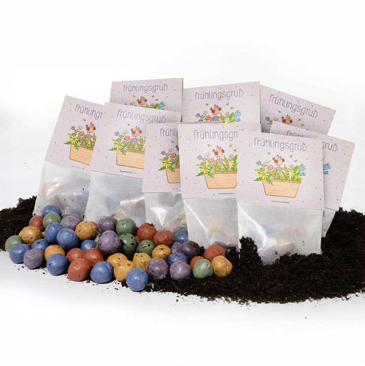 Sobre el suelo se encuentran varias bolsitas con la inscripción “Saludos de primavera” y decoradas con motivos florales, y delante de ellas hay bolas de semillas de colores.