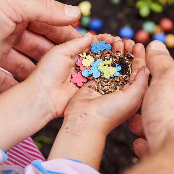 Las manos de los niños sostienen semillas de colores con formas de animales y flores sobre la tierra.